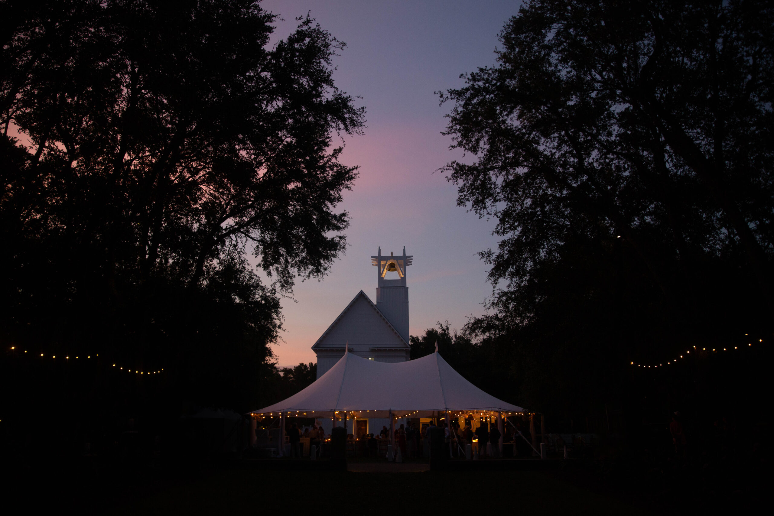 A tented wedding reception glowing as dusk ascends in the sky turns shades of pink and purple with a Seaside Chapel in the background during a destination Florida wedding