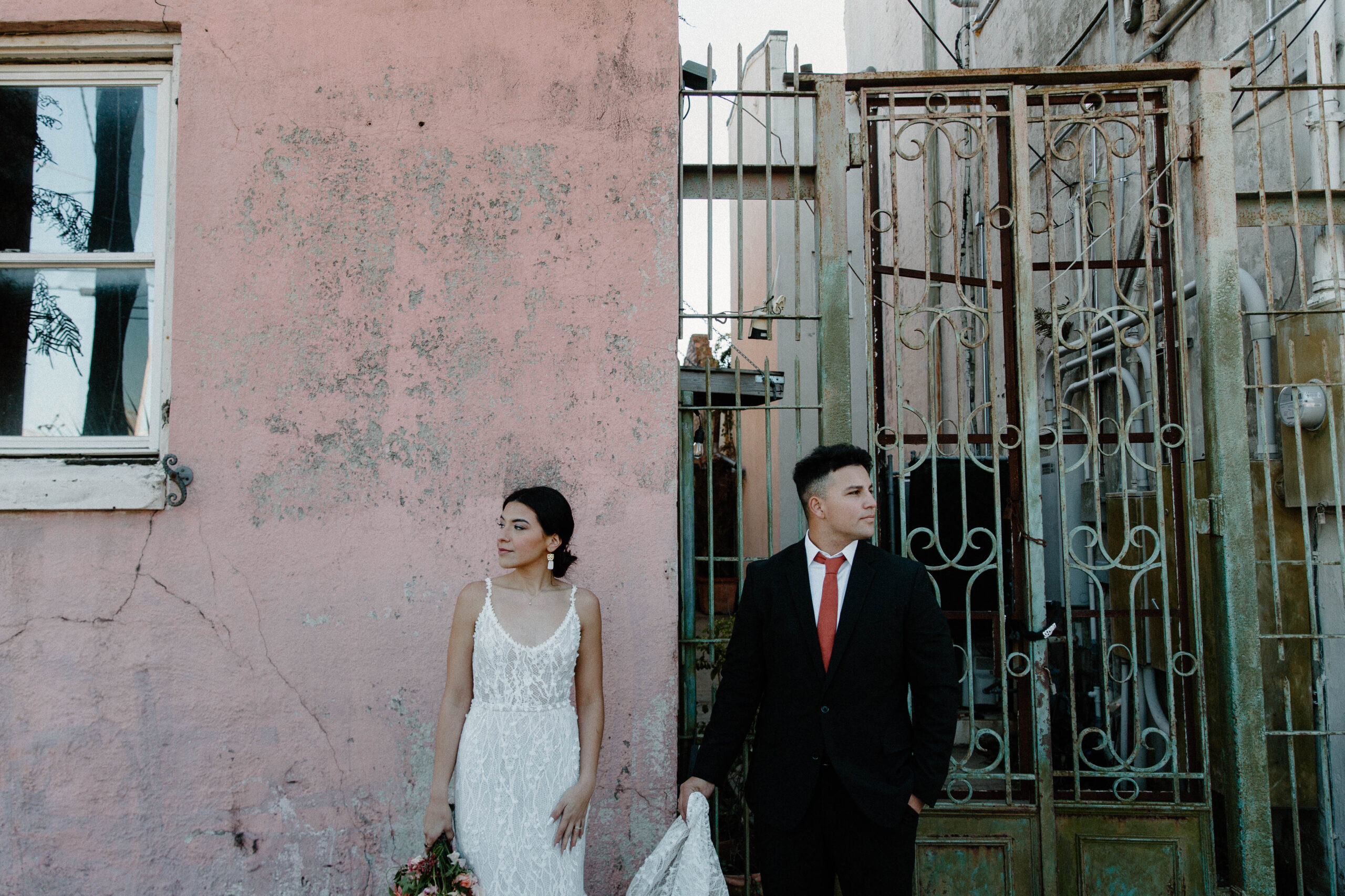A woman standing in front of a pink wall in a 3-D lace wedding dress as her partner stands next to her and hold the train of her dress during their urban elopement in New Orleans