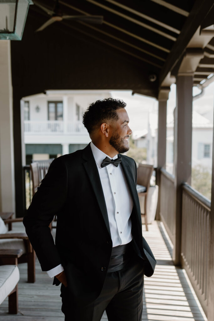 A groom standing on the second floor balcony with his hands in his pockets as he looks out over the town during their destinations luxury wedding on 30 A