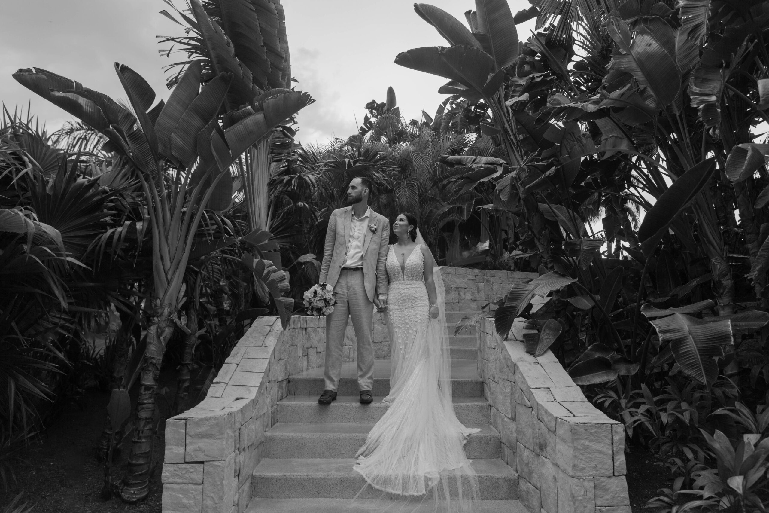 A couple in writing a tire standing on a set of steps, surrounded by tropical plants during their rainy, destinations wedding, a dream playa Mujeres resort in Cancun