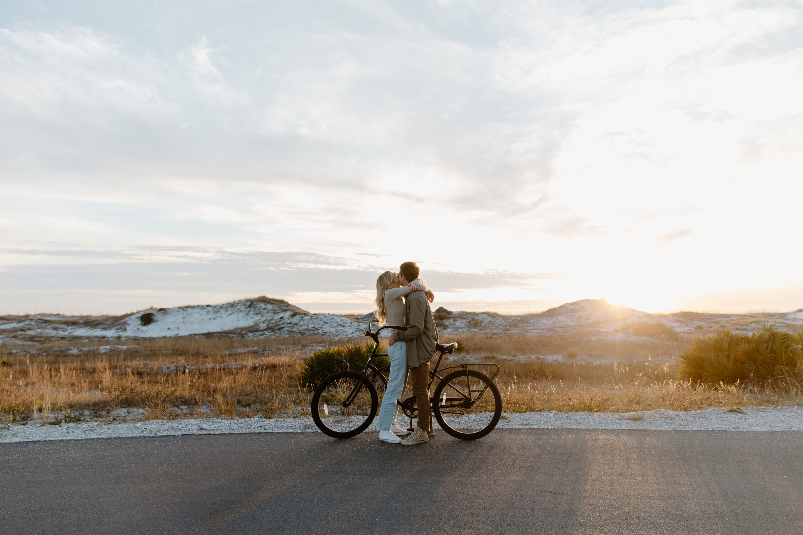 A couple standing together, straddling a bike as they look off at the sunset setting during their Florida state park. Couple photo shoot.