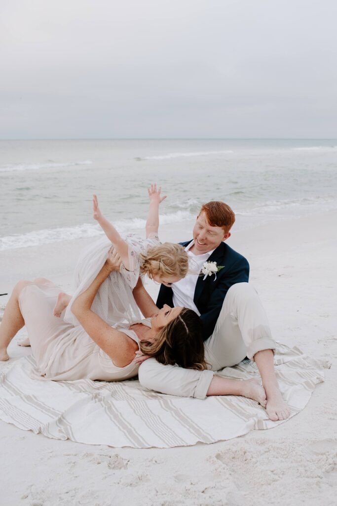 A newly wed couple with their child as the man is sitting on a blanket on the beach with his wife laying her head in his lap and her air planing their daughter as she laughs during their sunrise beach elopement in Santa Rosa Beach