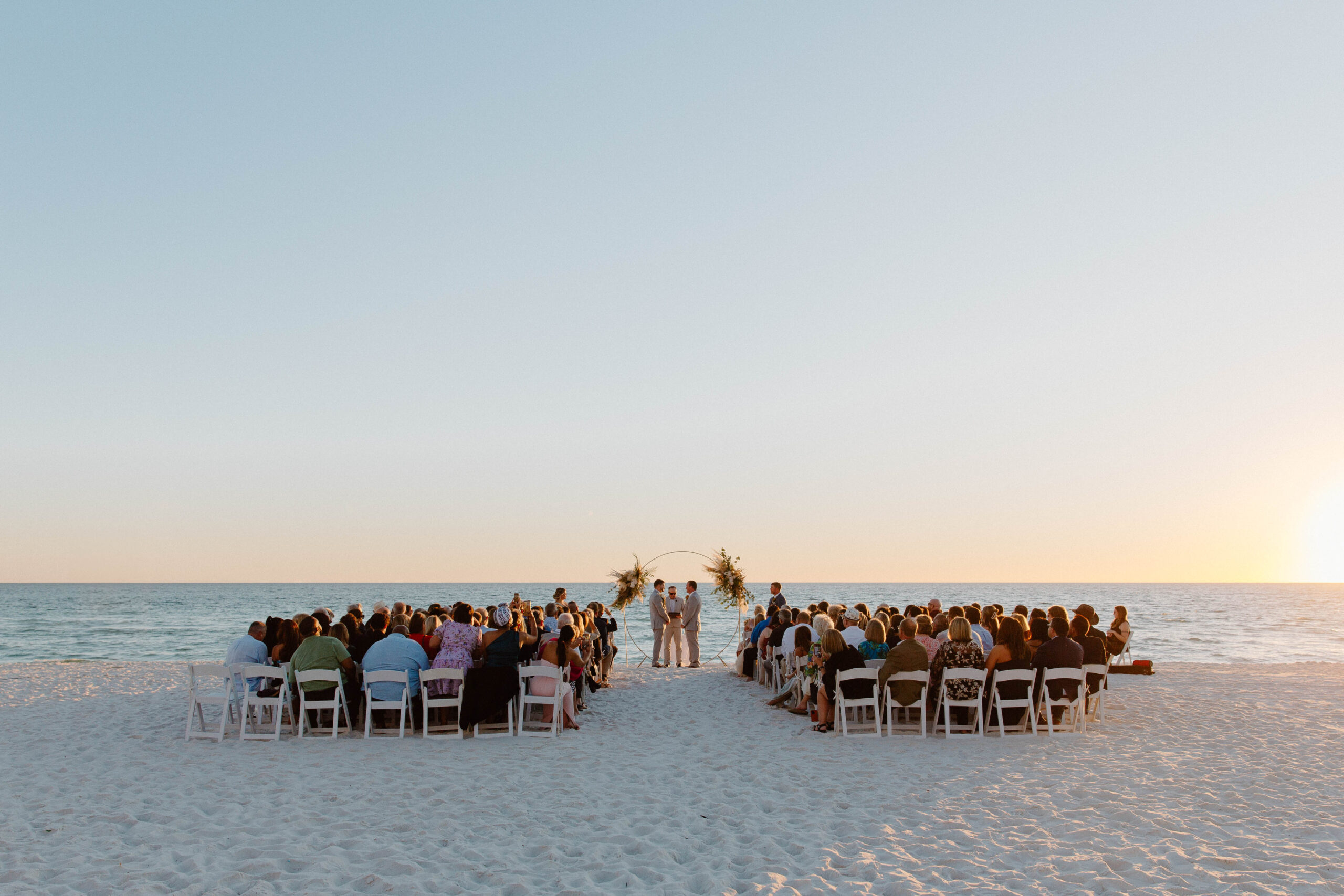 A man and his partner standing in front of a circular wedding, arch adorned with dried florals during their beach wedding ceremony in Florida