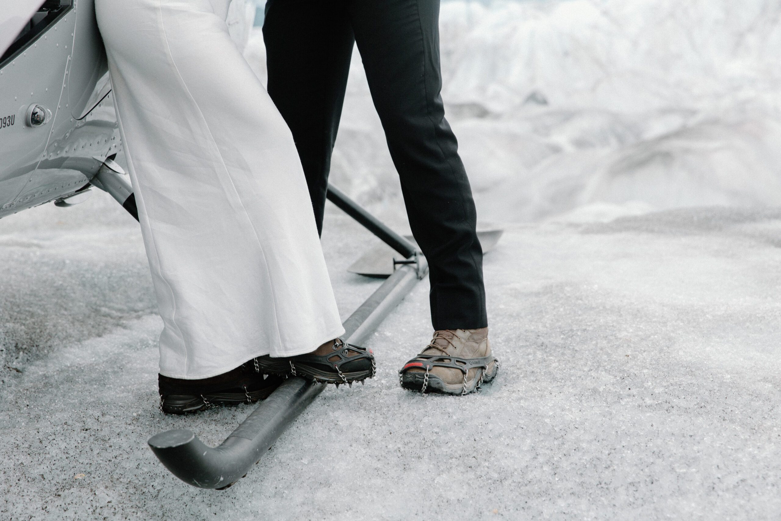 A couple standing on a glacier next to a helicopter with crampons on their hiking boots during their alaska helicopter elopement
