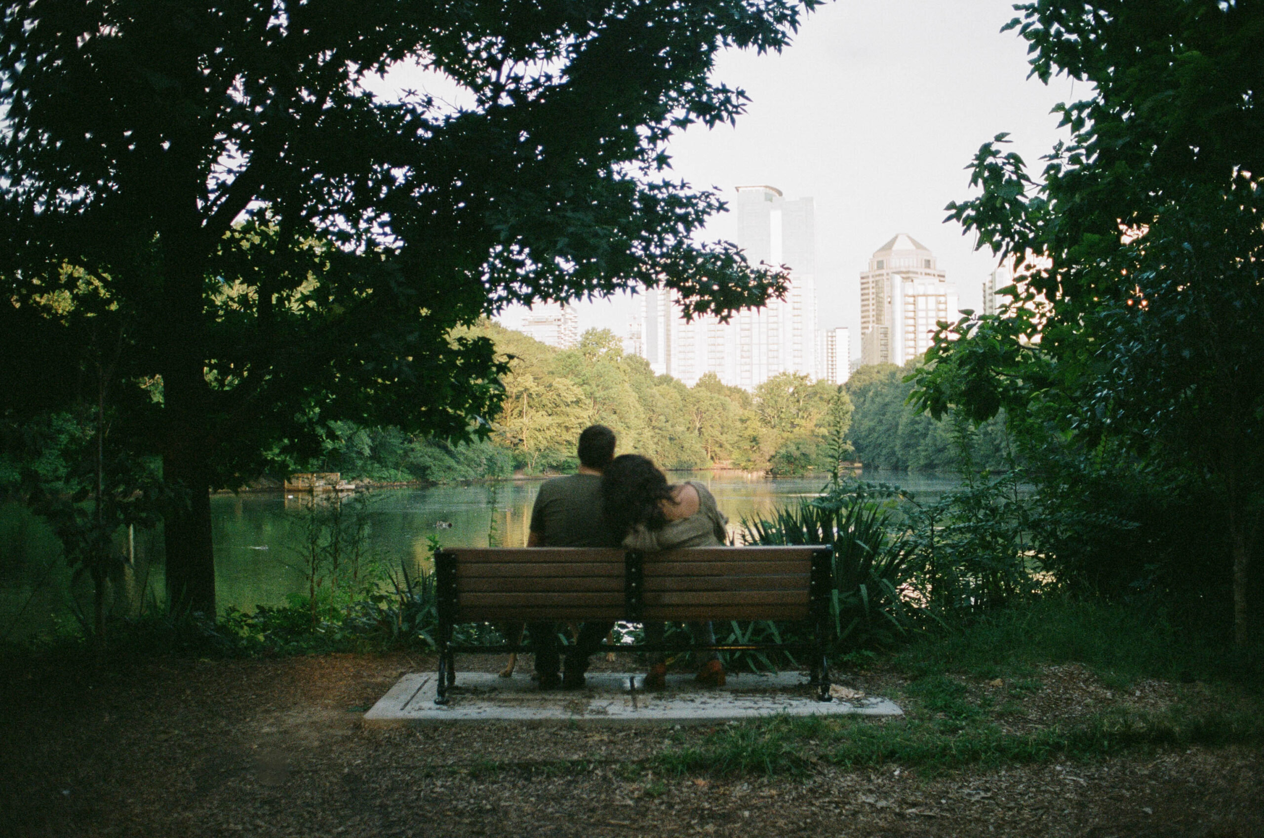 A couple sitting on a bench looking out over at the Atlanta skyline in the woman is resting her head on her partner shoulder during their Piedmont Park couple photo shoot