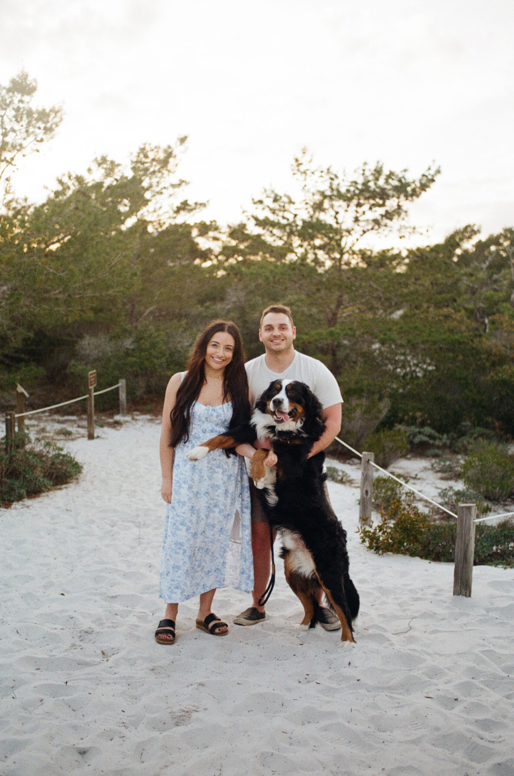 A couple standing and smiling as the man holds up their large St. Bernard dog, and the dog looks as if he is smiling as well during their Destin Florida photo shoot