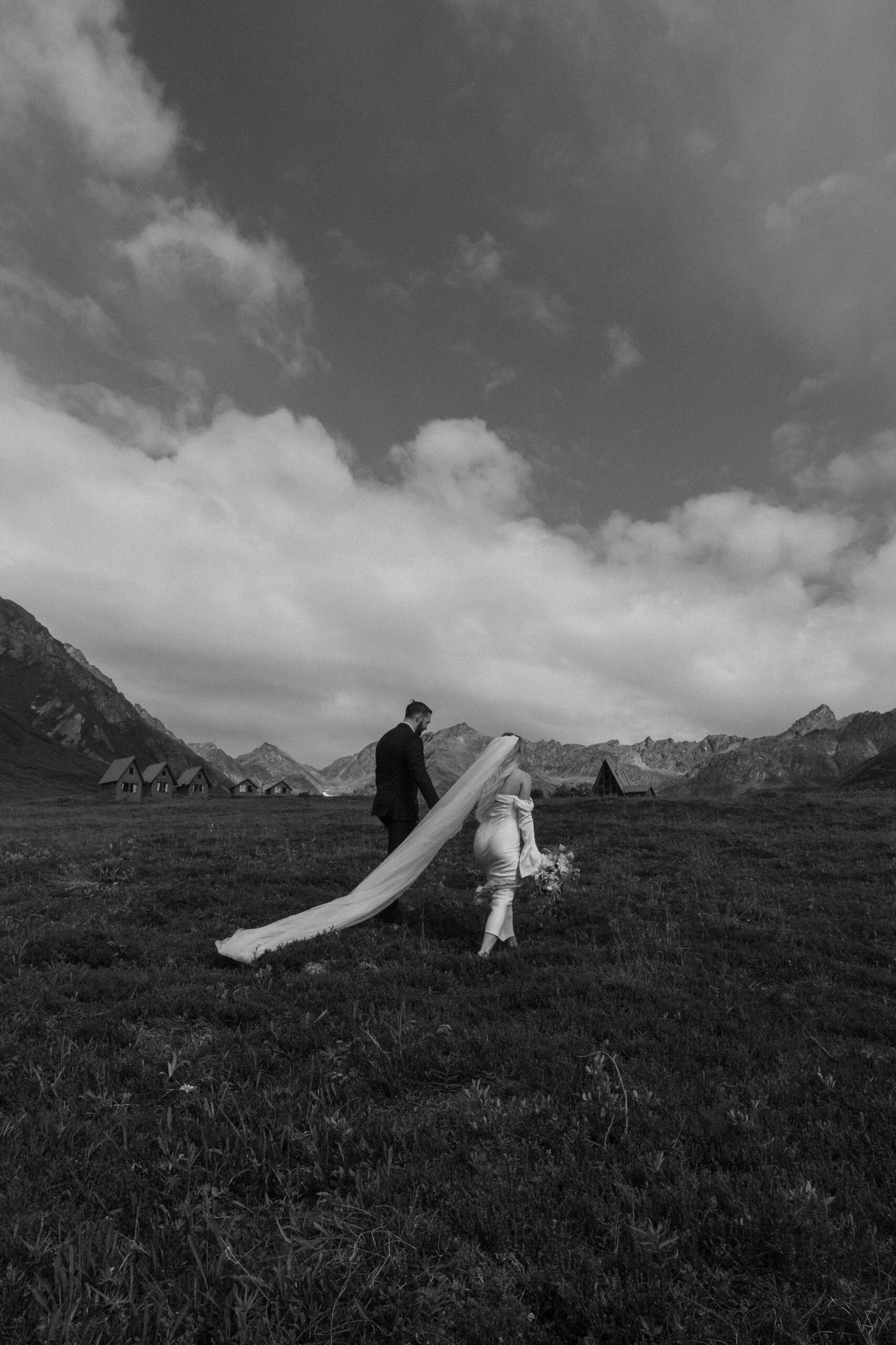 A man and a woman holding hands and walking up a hill as her veil trails behind them during their alaska elopement