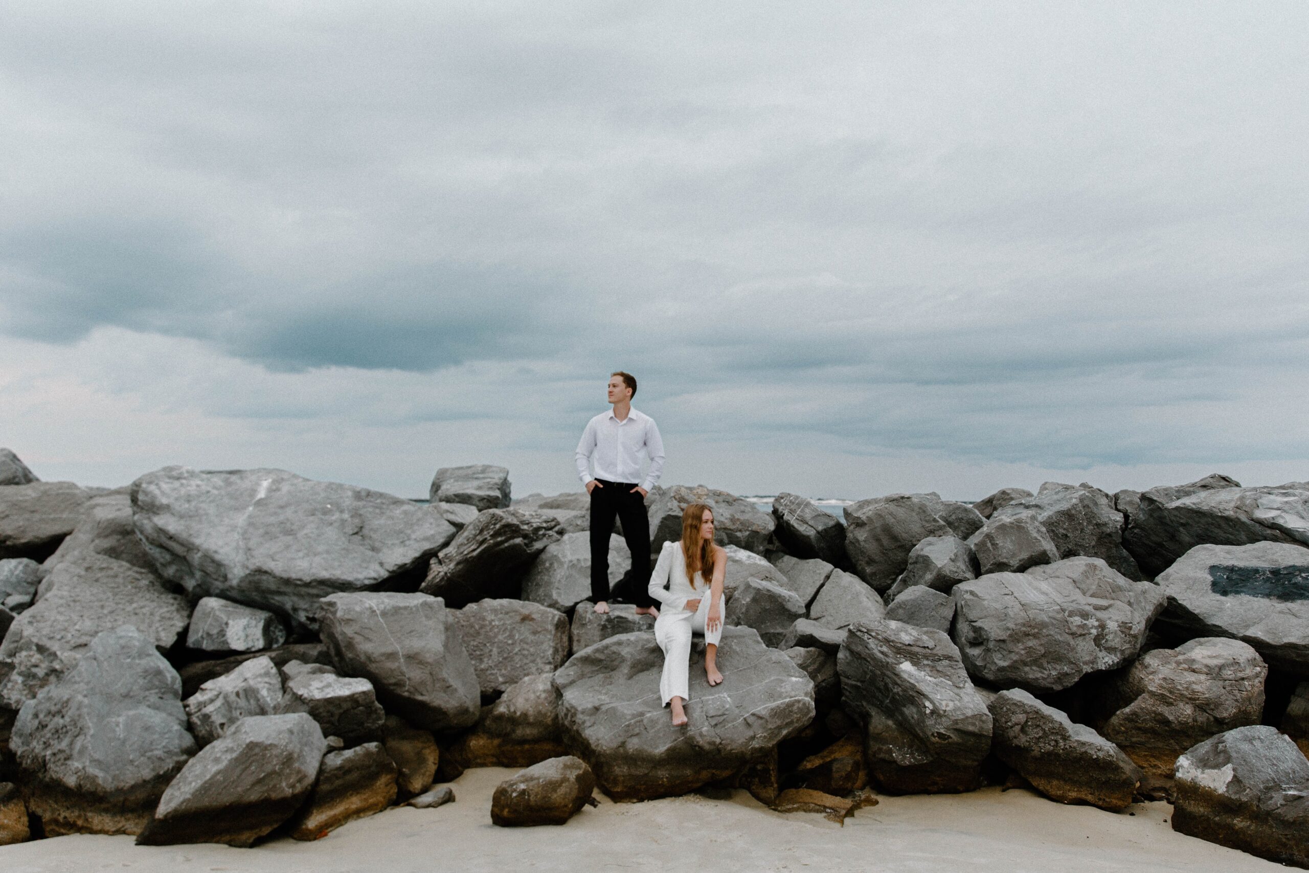 A man standing next to his partner with his hands in his pockets as she sits down on the rock during their beach engagement photos in Florida