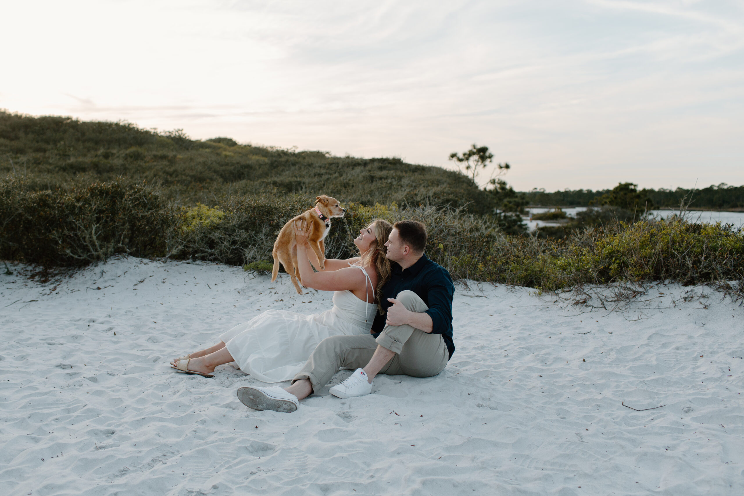 A woman and her partner sitting in the sand as the woman holds their puppy dog up during their Grayton Beach state park engagement photos