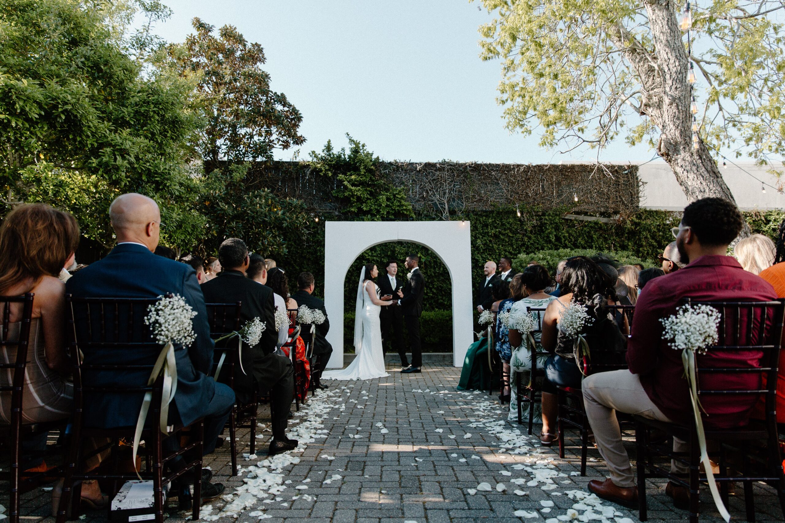 A couple holding hands as they stand in front of their white wedding arch and all their guests are sitting during their outdoor wedding ceremony in the courtyard at 5eleven Palafox