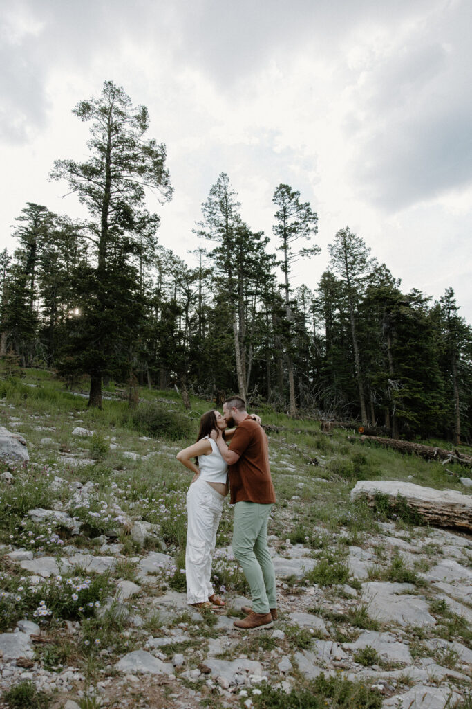 A man bringing his partner in for a kiss as she has her arms wrapped around him during their sunset couple session in the Sandia Mountains of New Mexico