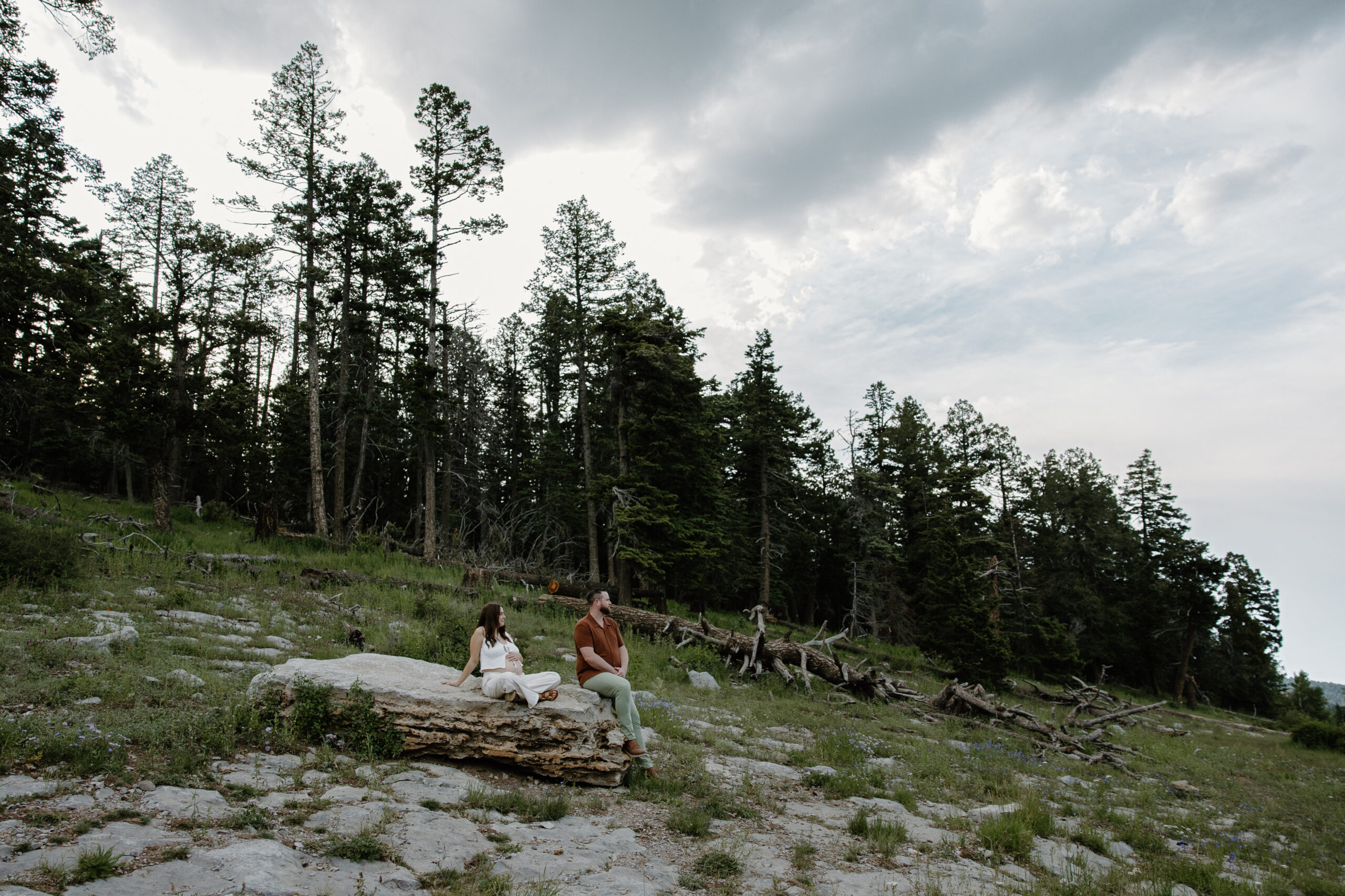 A woman sitting on a rock resting her hand on her belly as her partner sits on the edge looking off into the distance during their summer sunset photoshoot in the Sandia Mountains