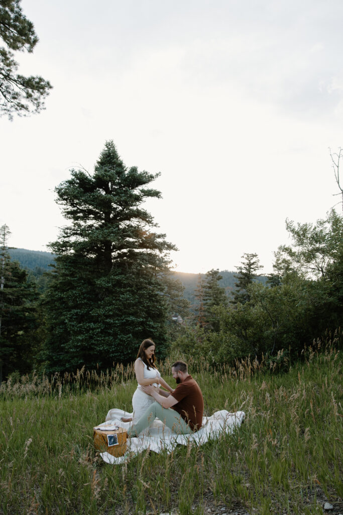 A woman kneeing in front of her partner as he sits on a picnic blanket and touches her growing belly during their summer maternity session in the Sandia Mountains