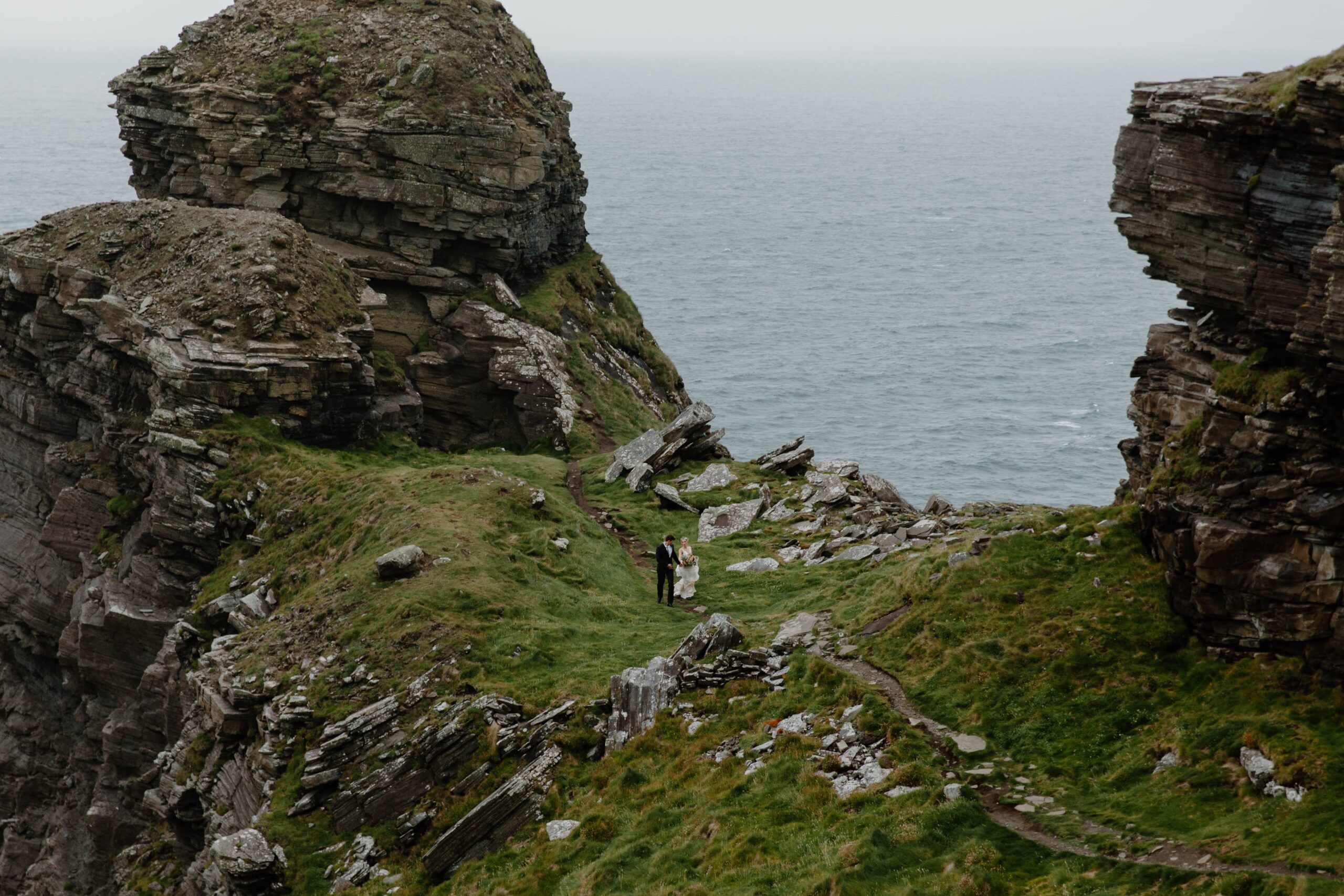 A couple in wedding attire holding hands and walking along a path surrounded by cliffs and water during their Cliffs of Moher elopement in Ireland