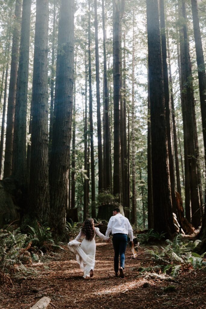 A couple in wedding attire holding hands and running through the forest during their Redwoods National Park elopement in California