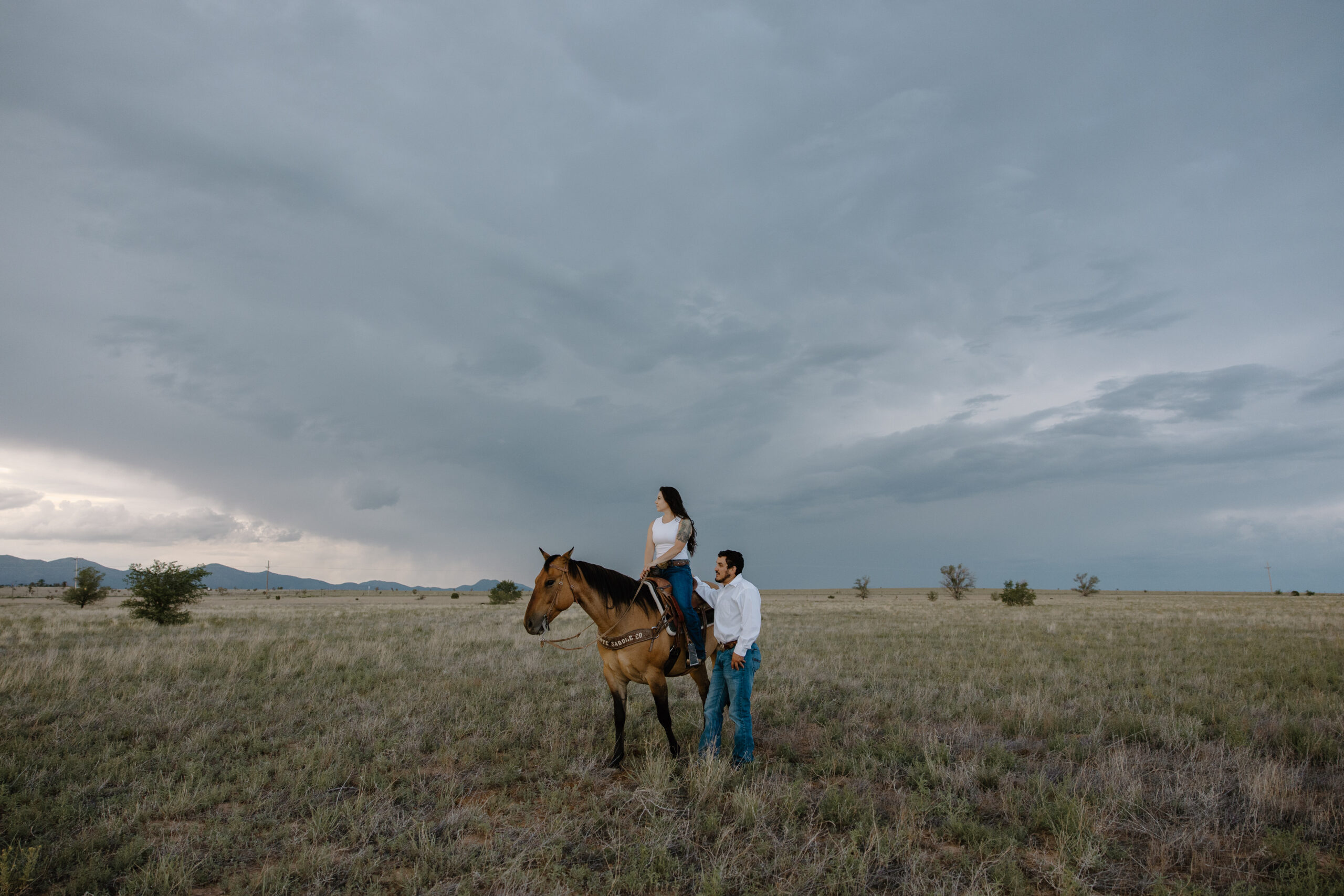 A woman sitting on her horse as her partner stands next to her and they look off into the distance during their Albuquerque couple photoshoot