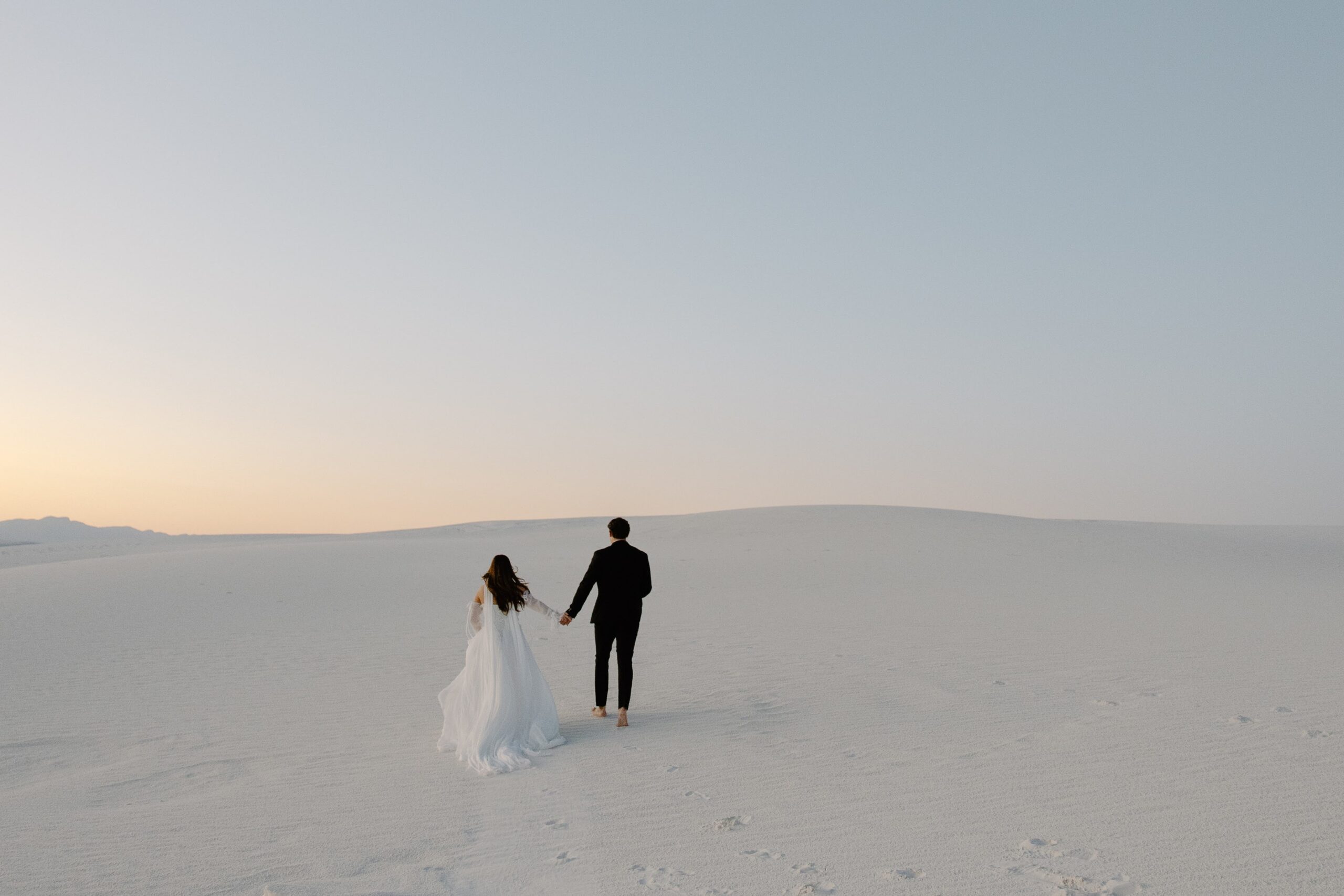 A couple holding hands and running over the sand dunes as the sun sets during their White Sands National Park wedding