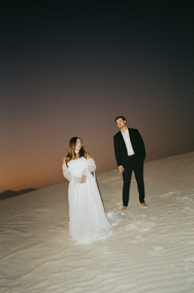 A couple in wedding attire standing apart from each other as the sun sets and paints the sky oranges during their White Sands National Park shot on 35mm film