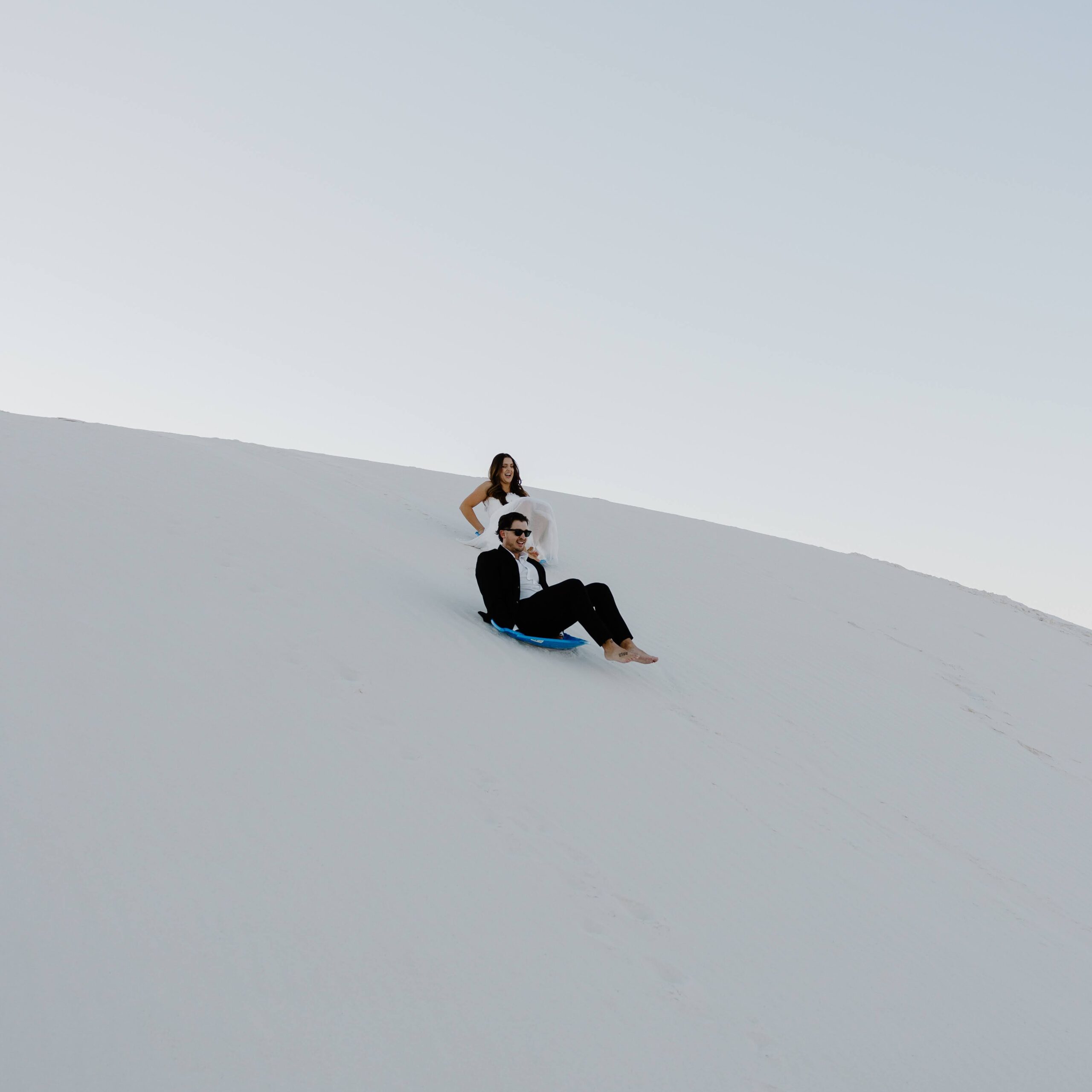 A couple in wedding attire sledding down sand dunes at White Sands National Park during a fall elopement