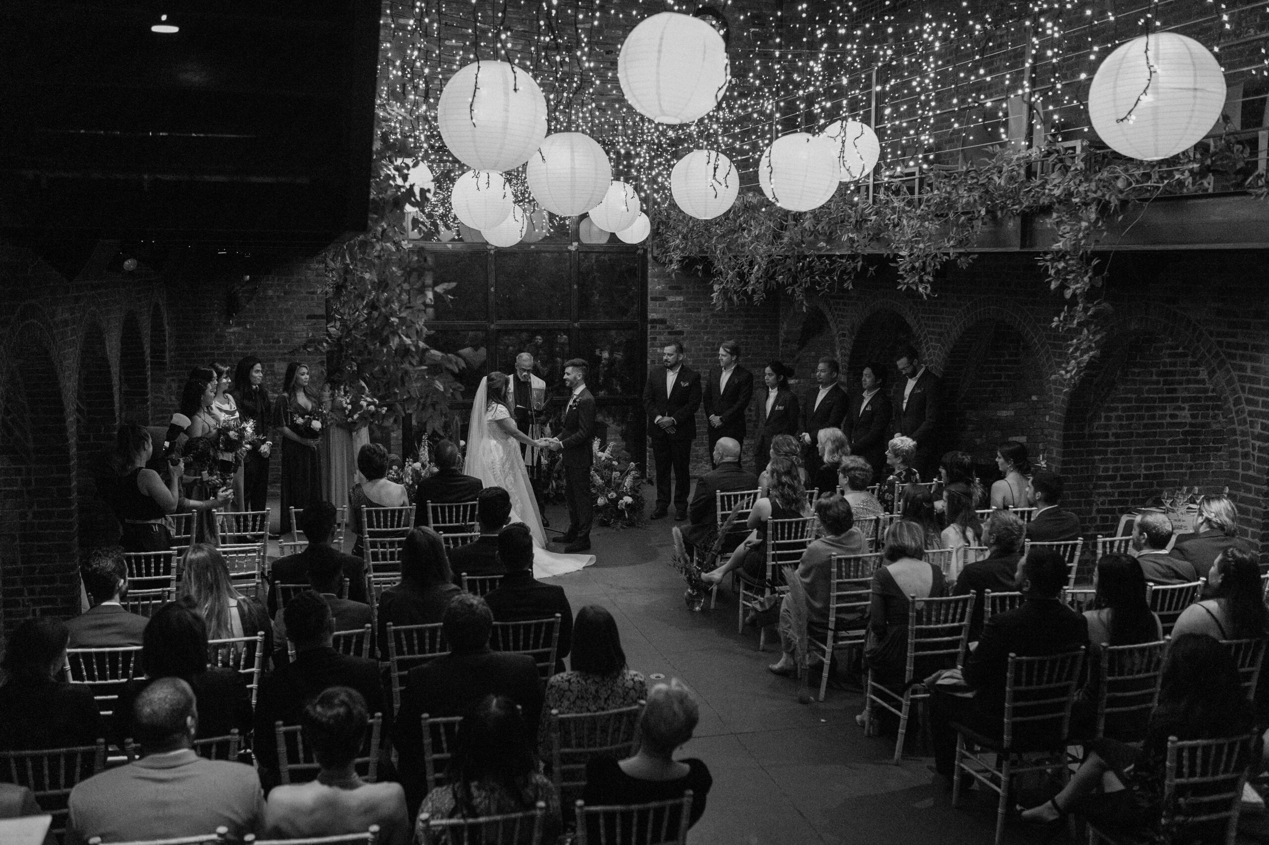 A couple in wedding attire standing up at the front of the aisle with their wedding party on either side of them as they say vows during their fall wedding in NYC