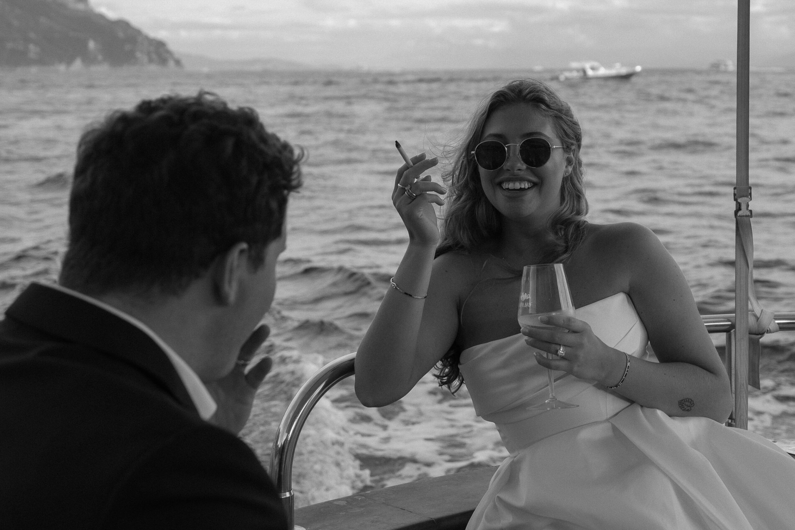 A woman in a wedding dress laughing as she smokes a cigarette and drinks wine on a boat on the Mediterranean Sea during her all day adventure elopement on the Amalfi Coast