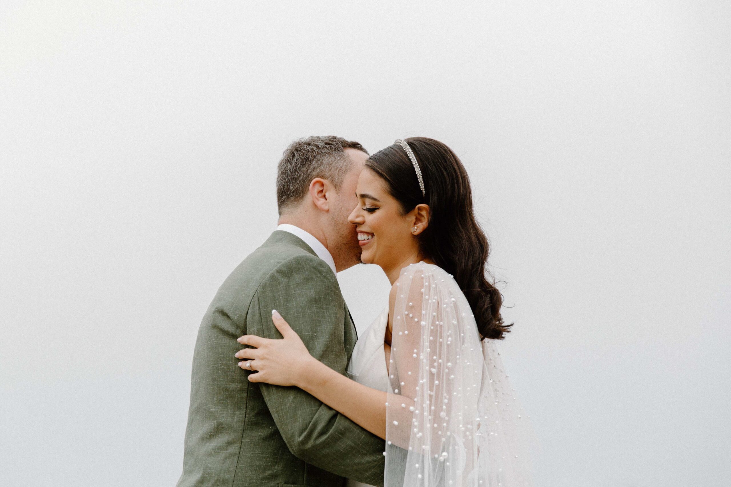 A man whispering into his partners ear as she laughs and holds on to him during their fall wedding