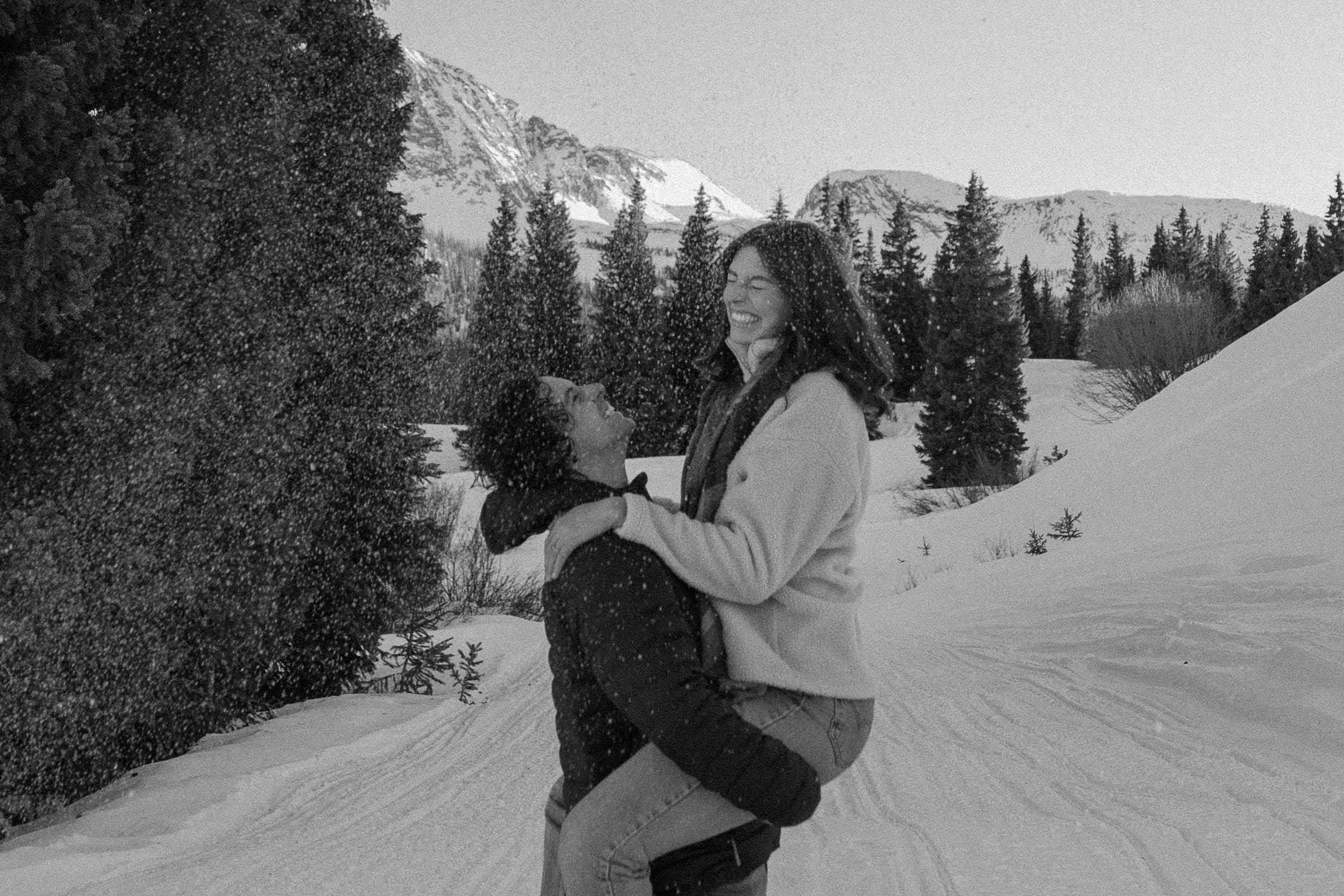 A woman and her partner laughing as snow falls around them during their sunset photoshoot in the mountains of Colorado