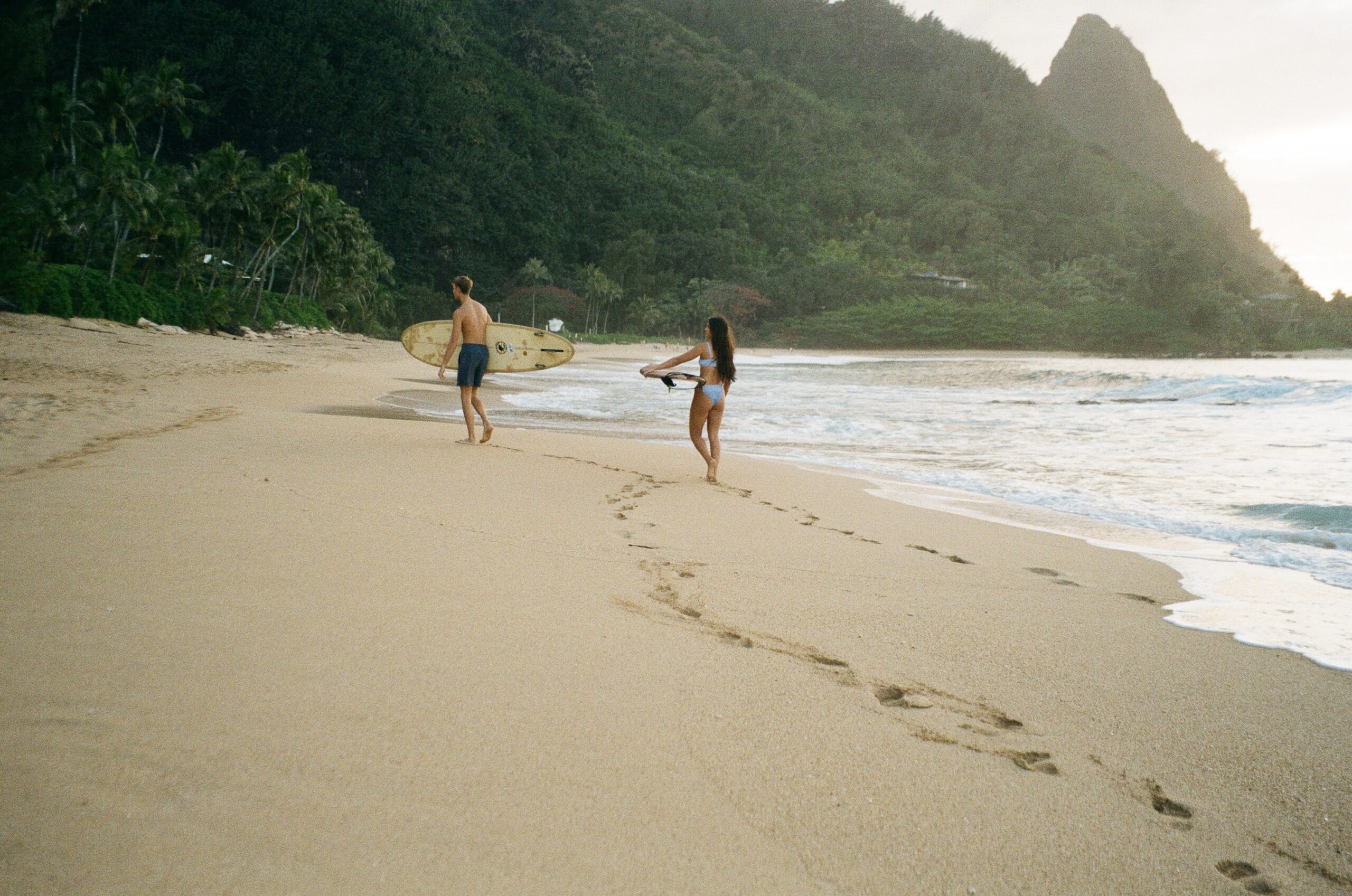 A couples foot prints in the sand as they walk far down the beach, holding surfboards captured on 35 mm film in Hawaii