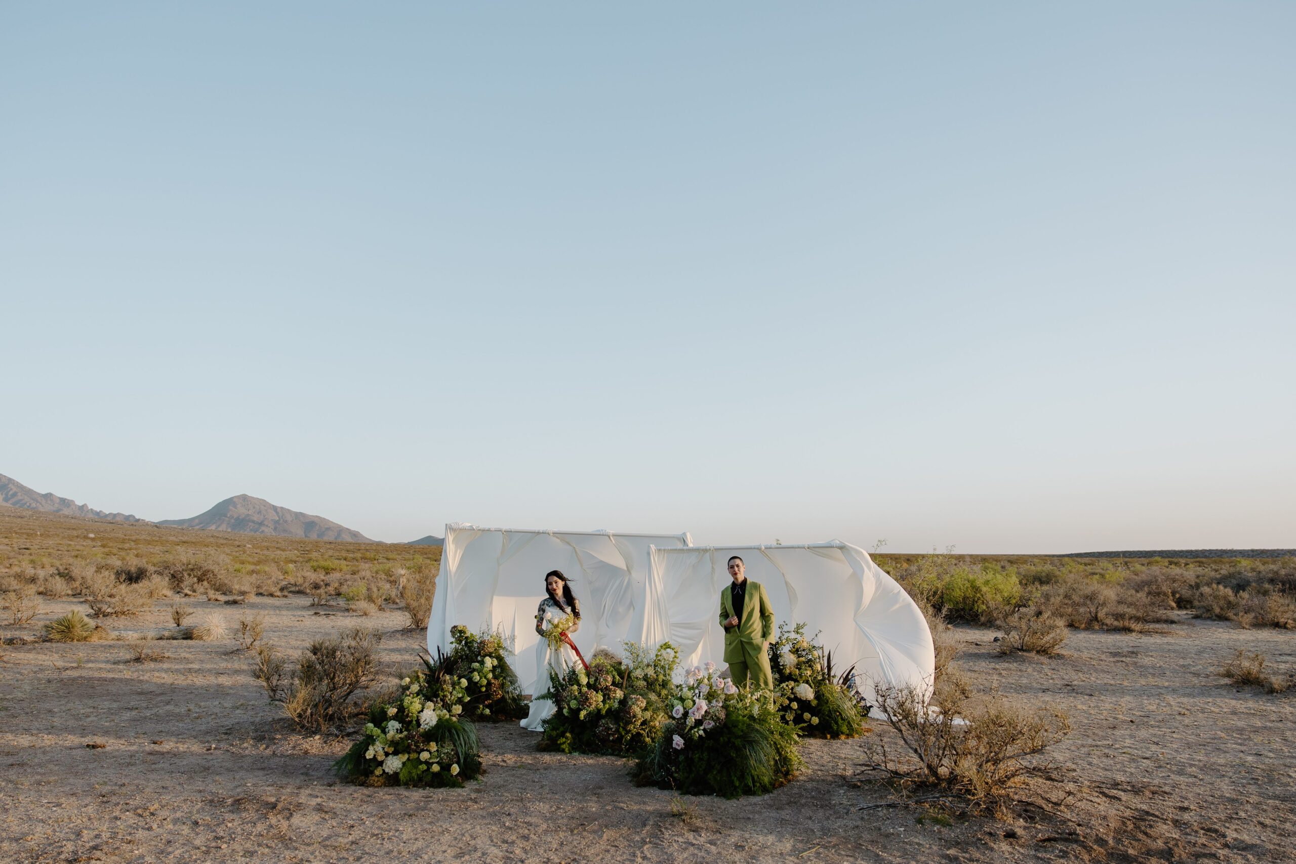 A couple and wedding attire standing apart from each other in front of a large drapery, and among florals during their desert wedding out west