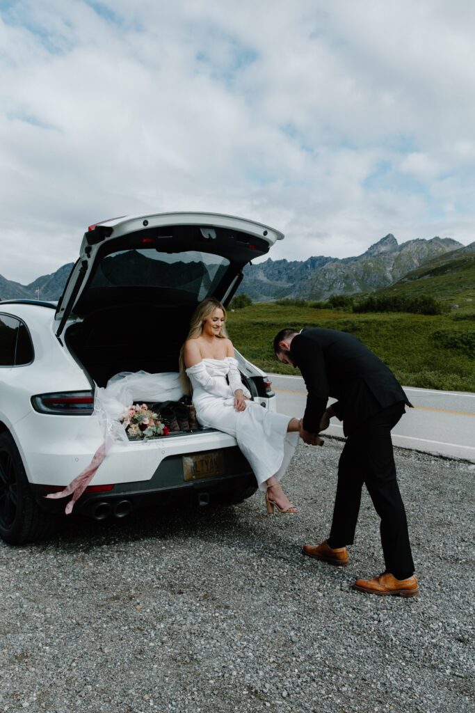 A woman sitting in the back of a car as her partner helps her get her heel on during their all day adventure elopement in Alaska