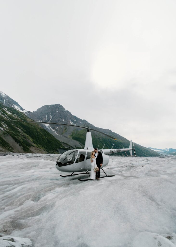 Couple in wedding attire standing in front of a helicopter after taking a ride in it out to a glacier during their elopement in Alaska