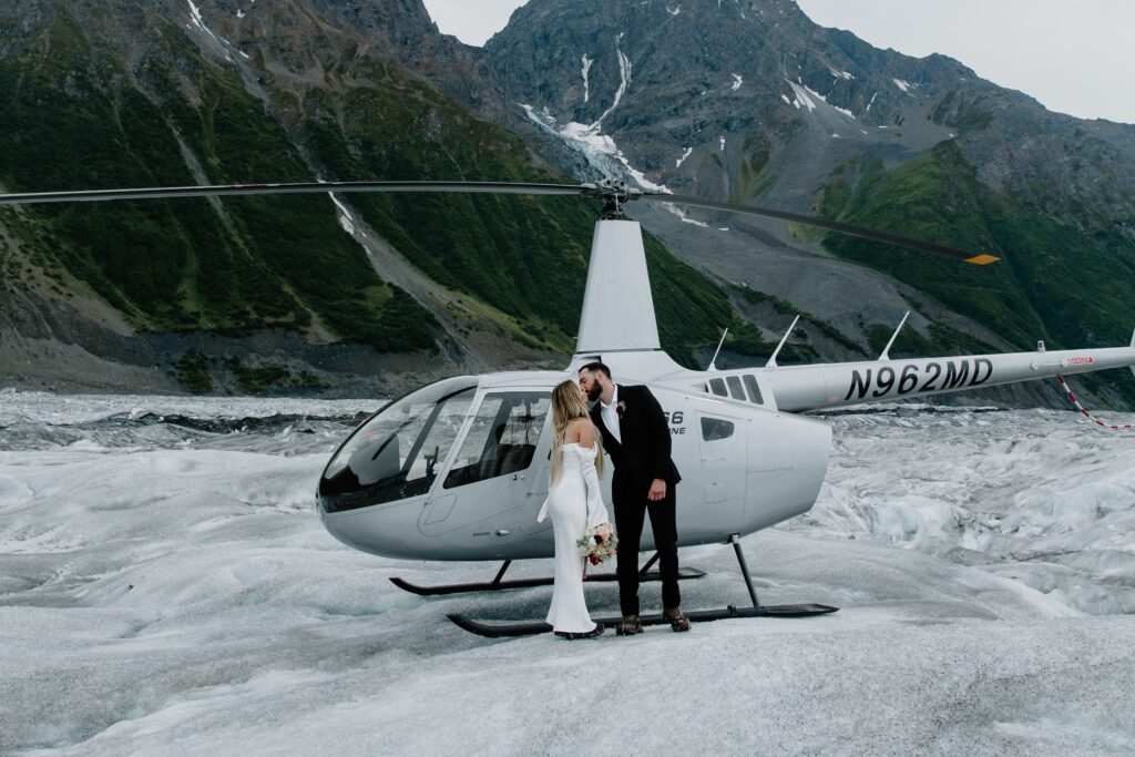 A couple in wedding attire sharing a kiss in front of a helicopter which was one of their adventure elopement ideas in Alaska