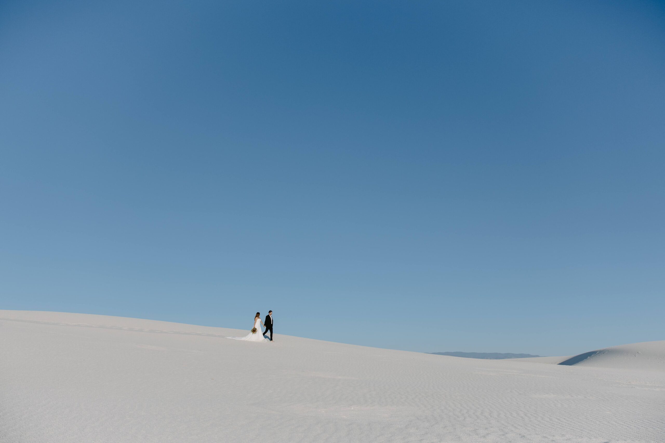 A couple holding hands and walking along the top of a sand dune during their New Mexico fall wedding
