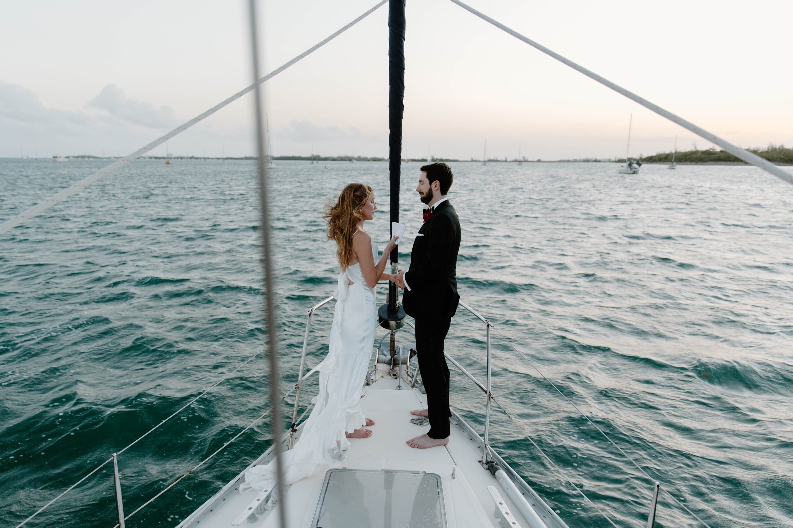 A couple in wedding attire at the front of a sailboat, reading their vows to each other as the sun rises over the Florida Keys during their key west elopement