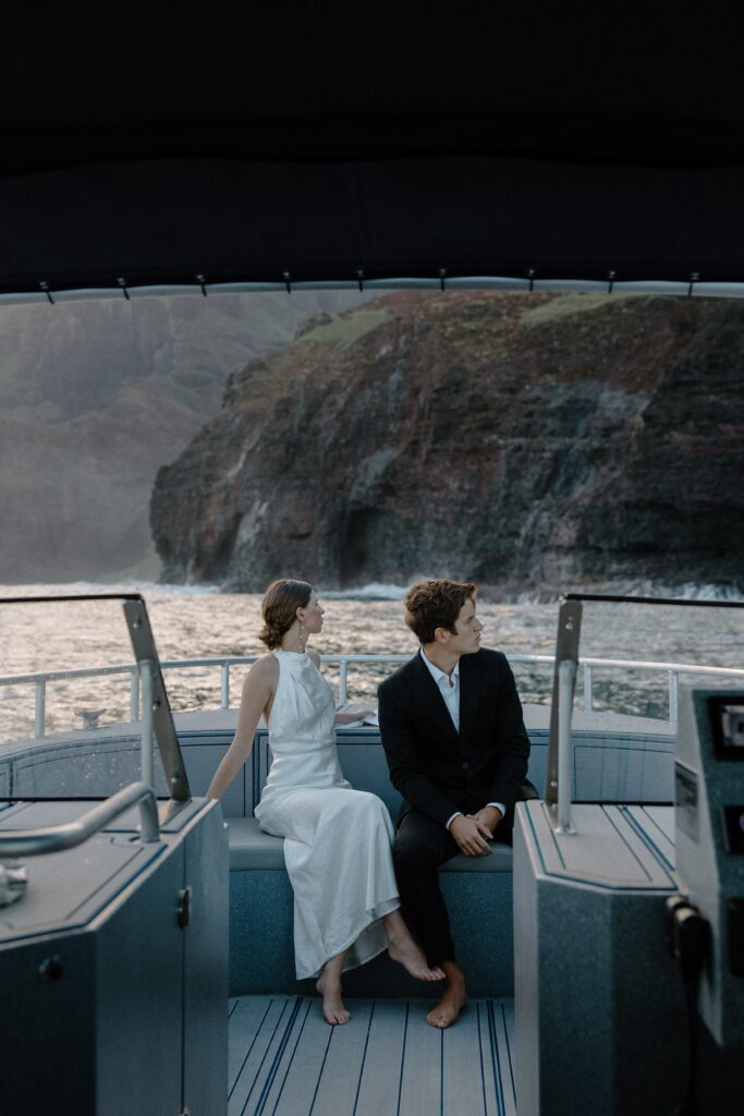 A couple sitting at the front of a boat as it goes along the Nā Pali coast during their spring adventure sunrise elopement Kauai boat elopement
