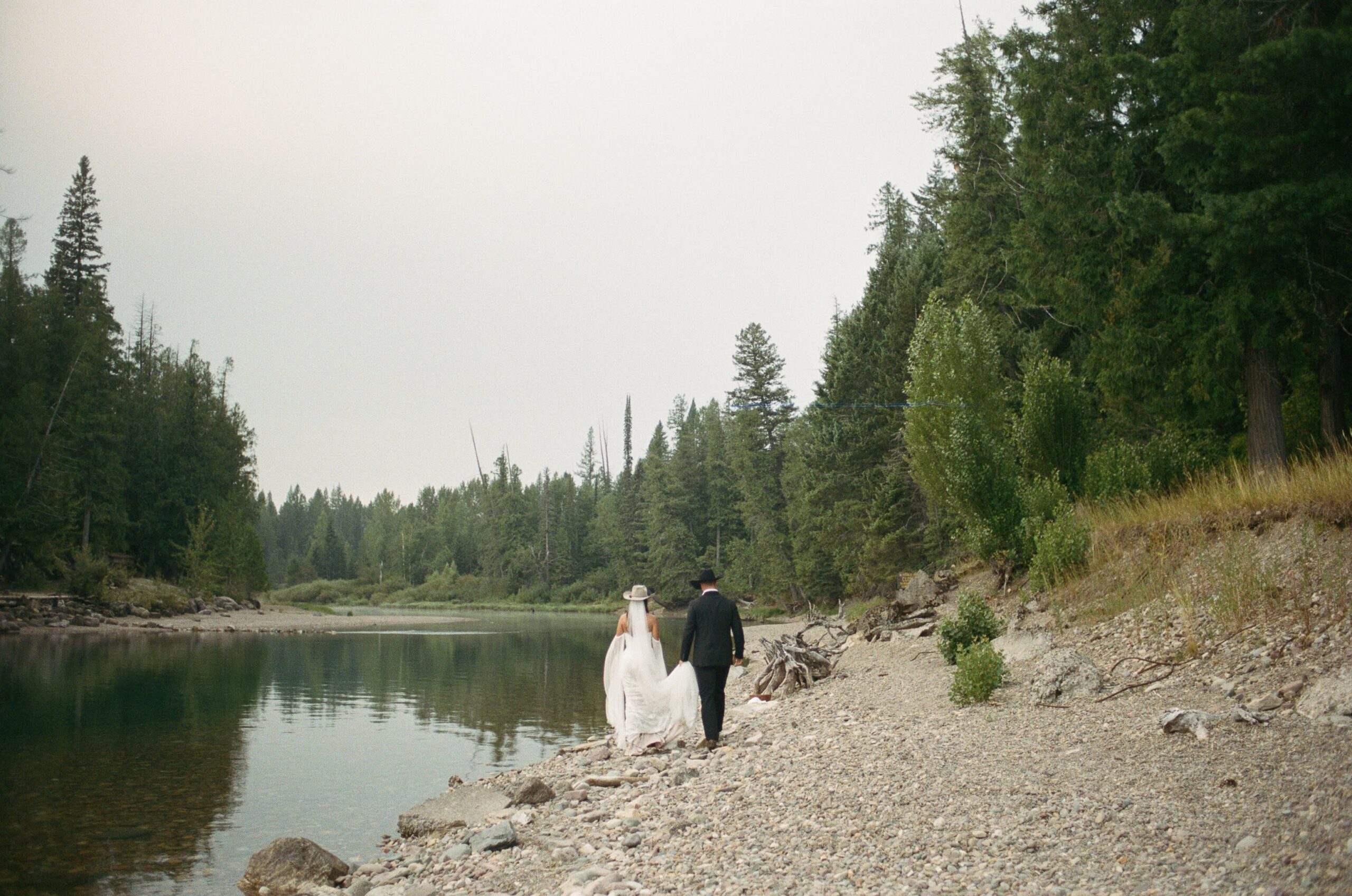 A couple in wedding attire holding hands and walking along the lakes edge during their all day elopement in Glacier National Park