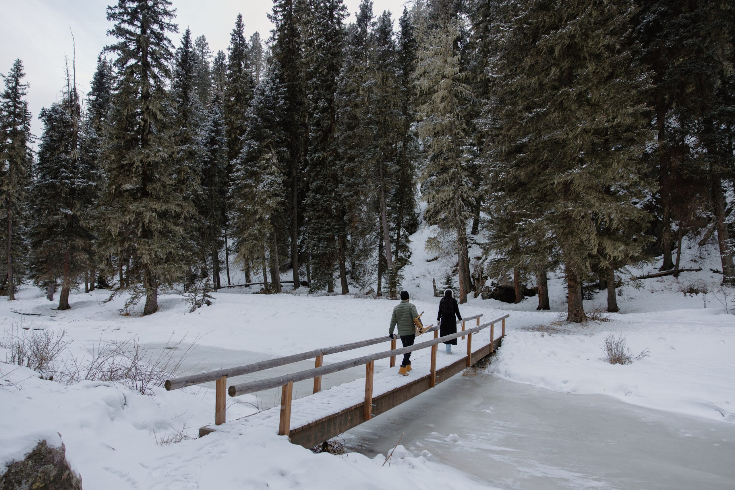 A couple in winter attire walking across a bridge over a frozen river during their winter photoshoot in New Mexico