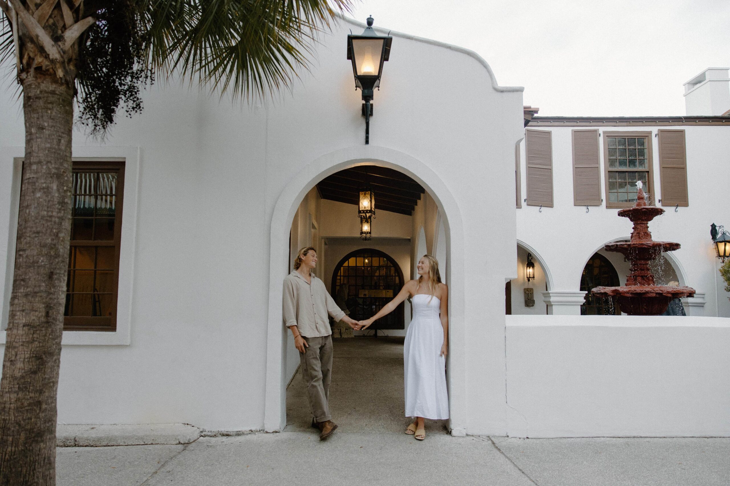 A couple standing on opposite sides of a door frame as they hold hands in the middle and smile at each other
