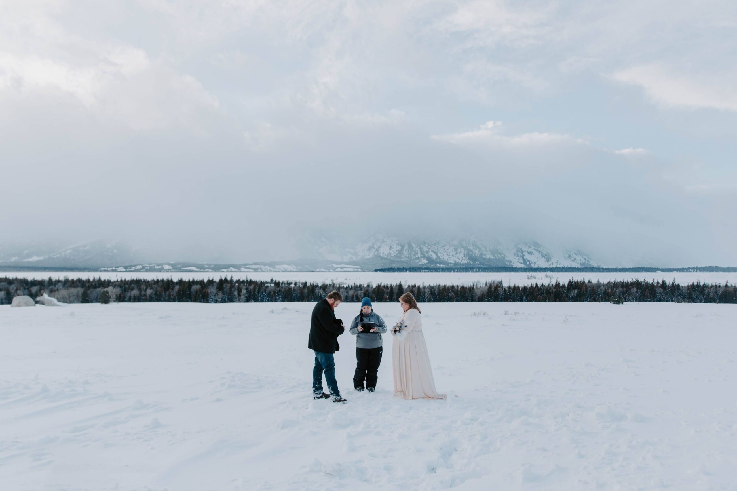 A woman and her partner standing in the snow with their officiant saying vows to each other with the Grand Tetons in the background during their Wyoming wedding