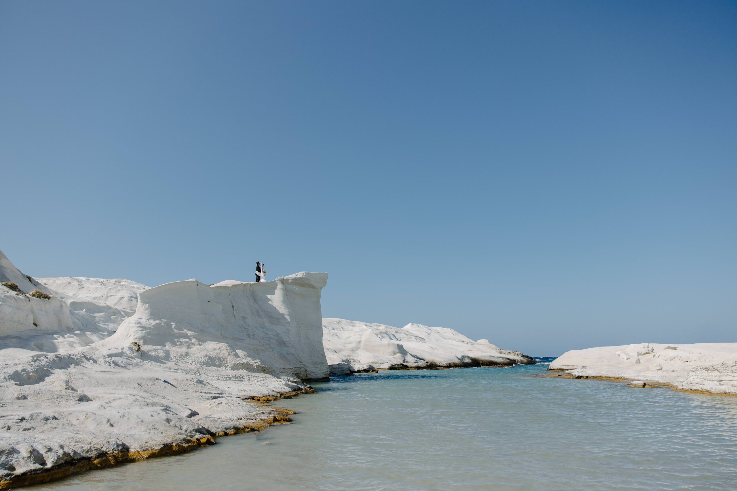 White volcanic cliffs meeting the sea in Milos, Greece with the couple up on a rock formation high above the water
