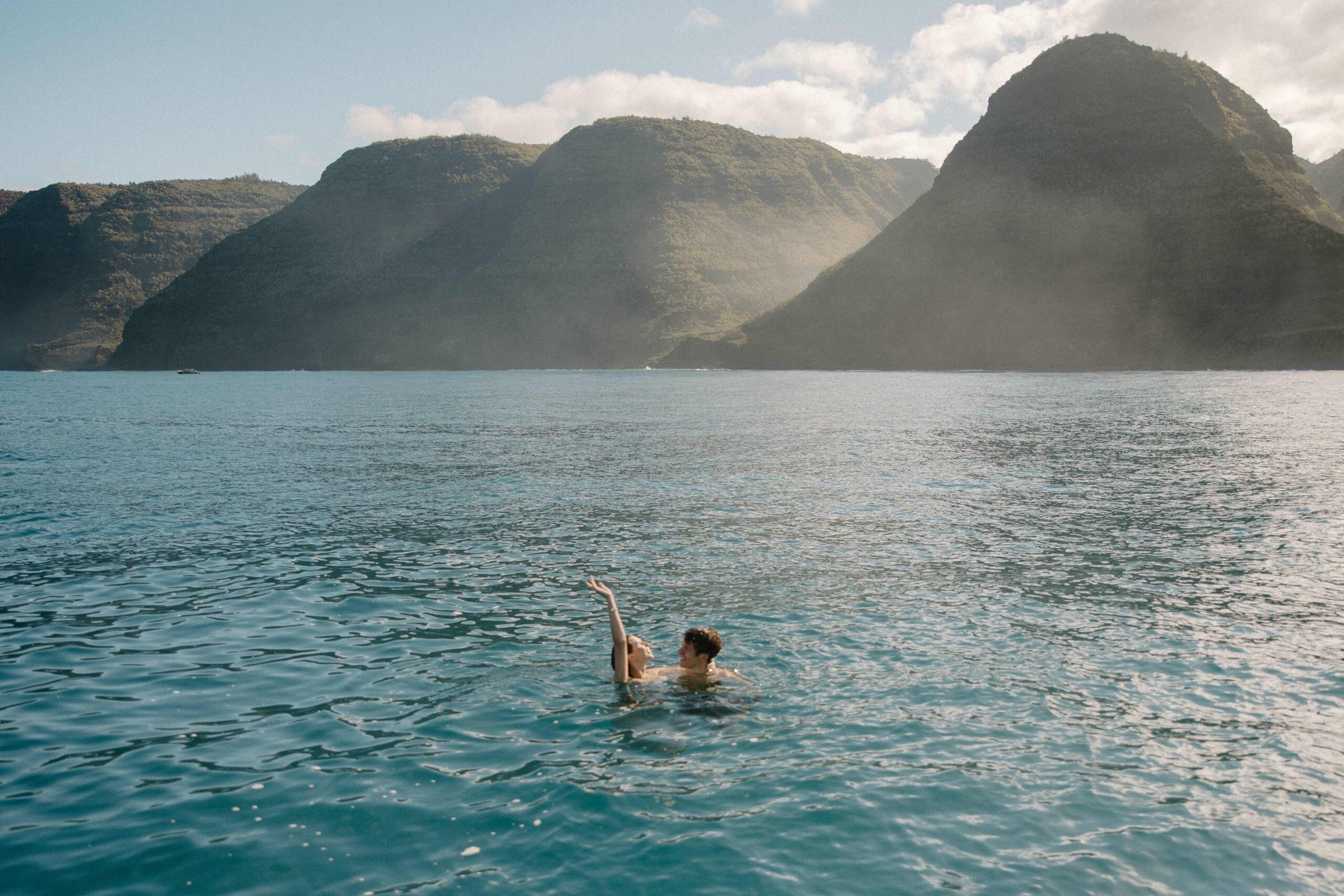 A woman and her partner swimming in the ocean as she throws her arm up during their Nā Pali Coast sunrise wedding