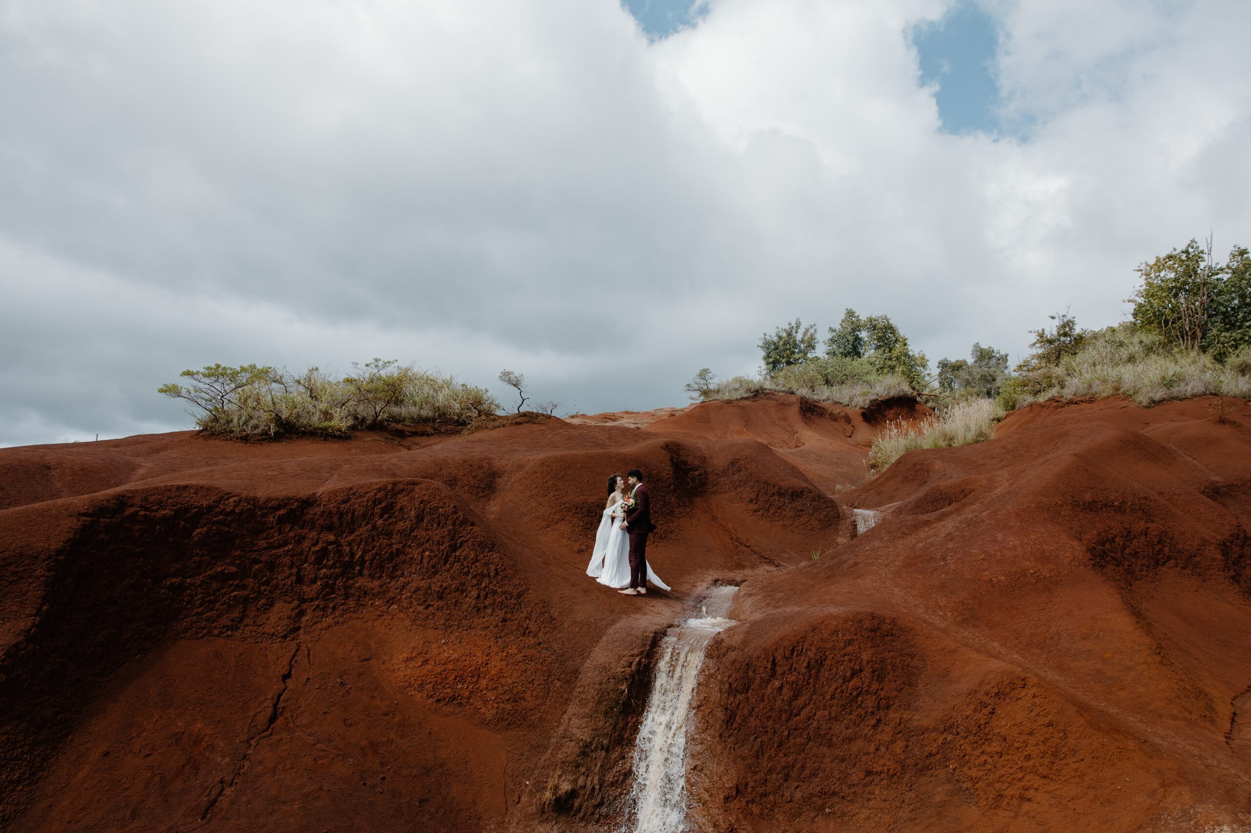 Wide view of bride and groom on red cliffs with waterfall scenery in Kauaʻi