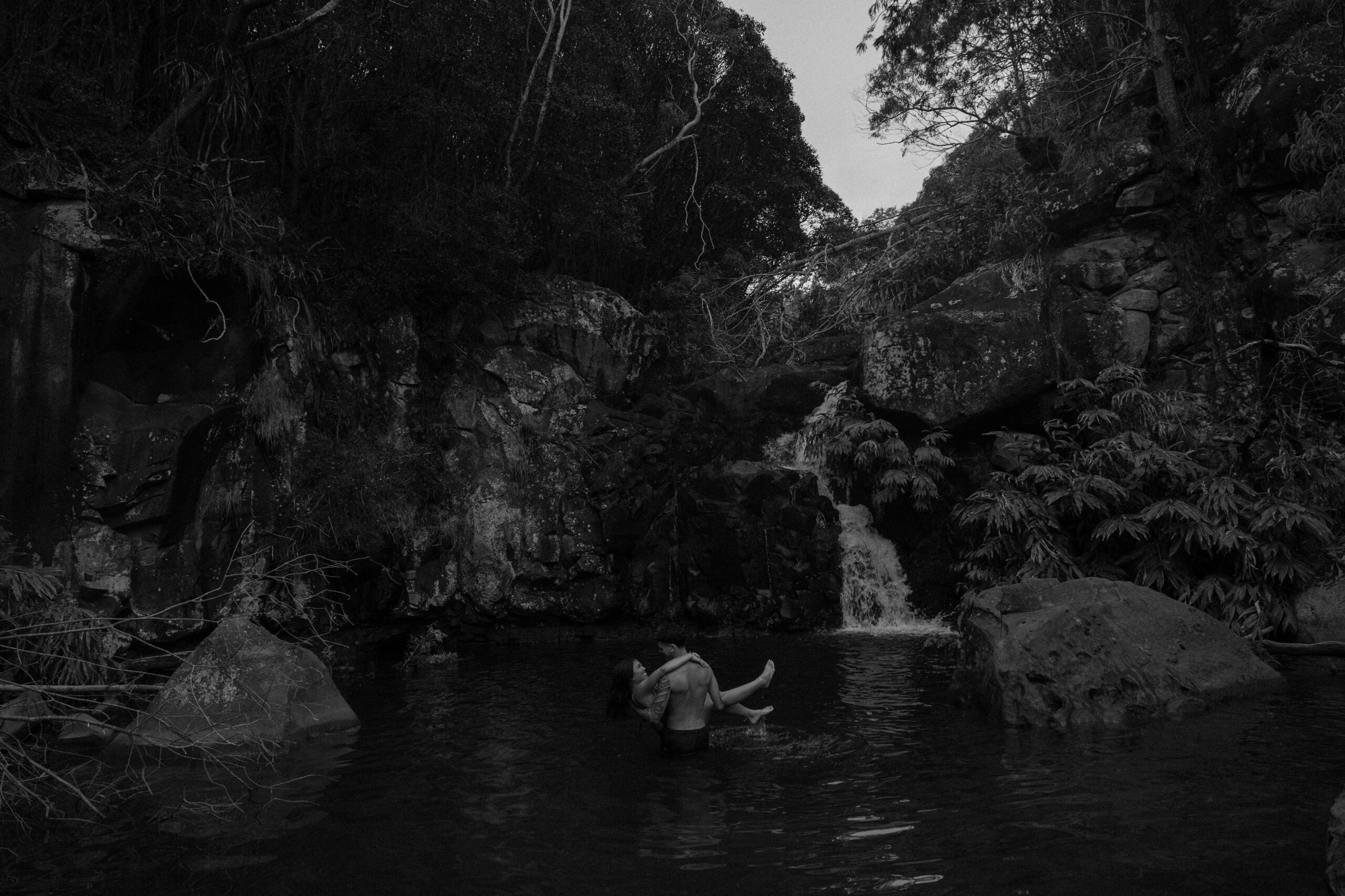 Man holding his partner as they are swimming beneath a hidden waterfall surrounded by lush canyon cliffs in Waimea Canyon State Park.