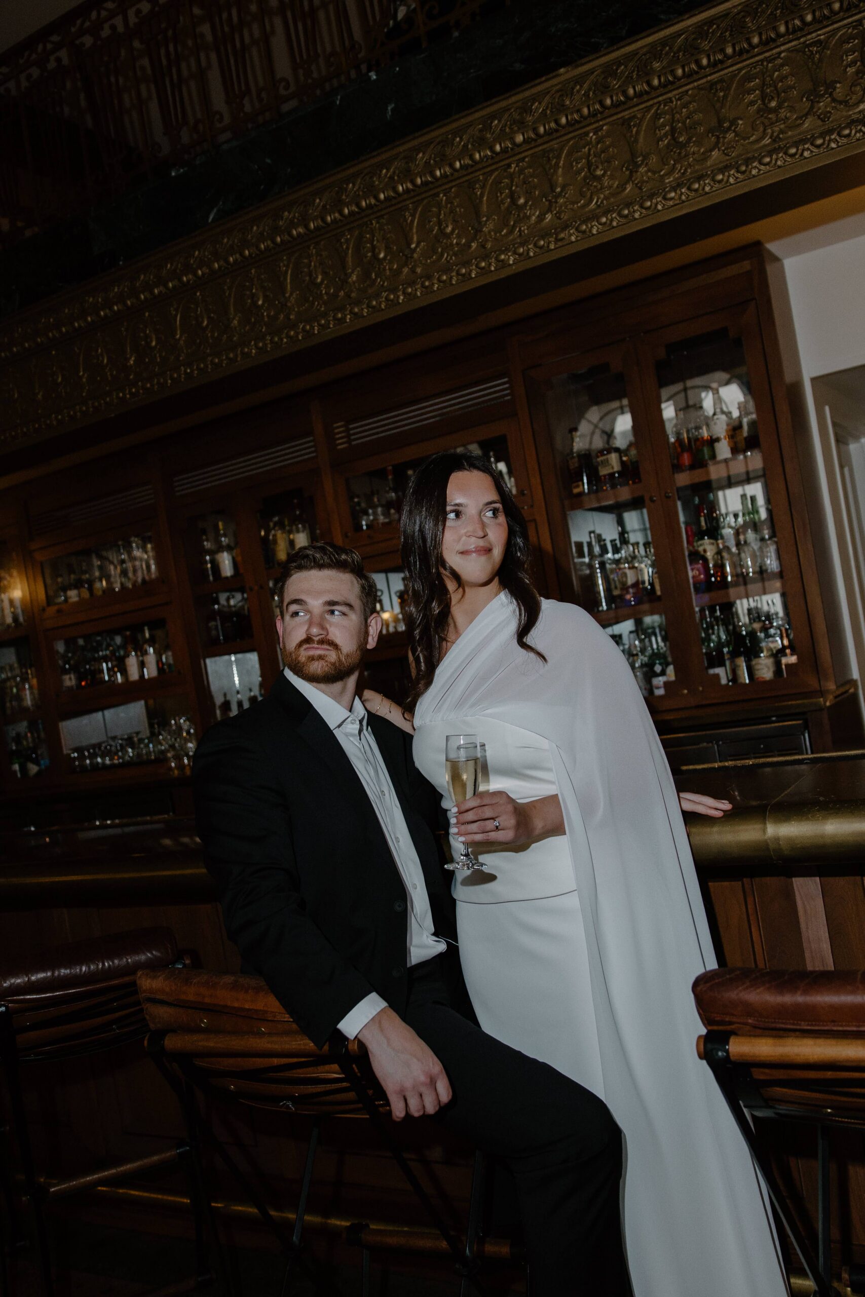 A man is sitting at a bar with his partner standing next to him as she's holding a glass of champagne during their downtown Nashville engagement photos