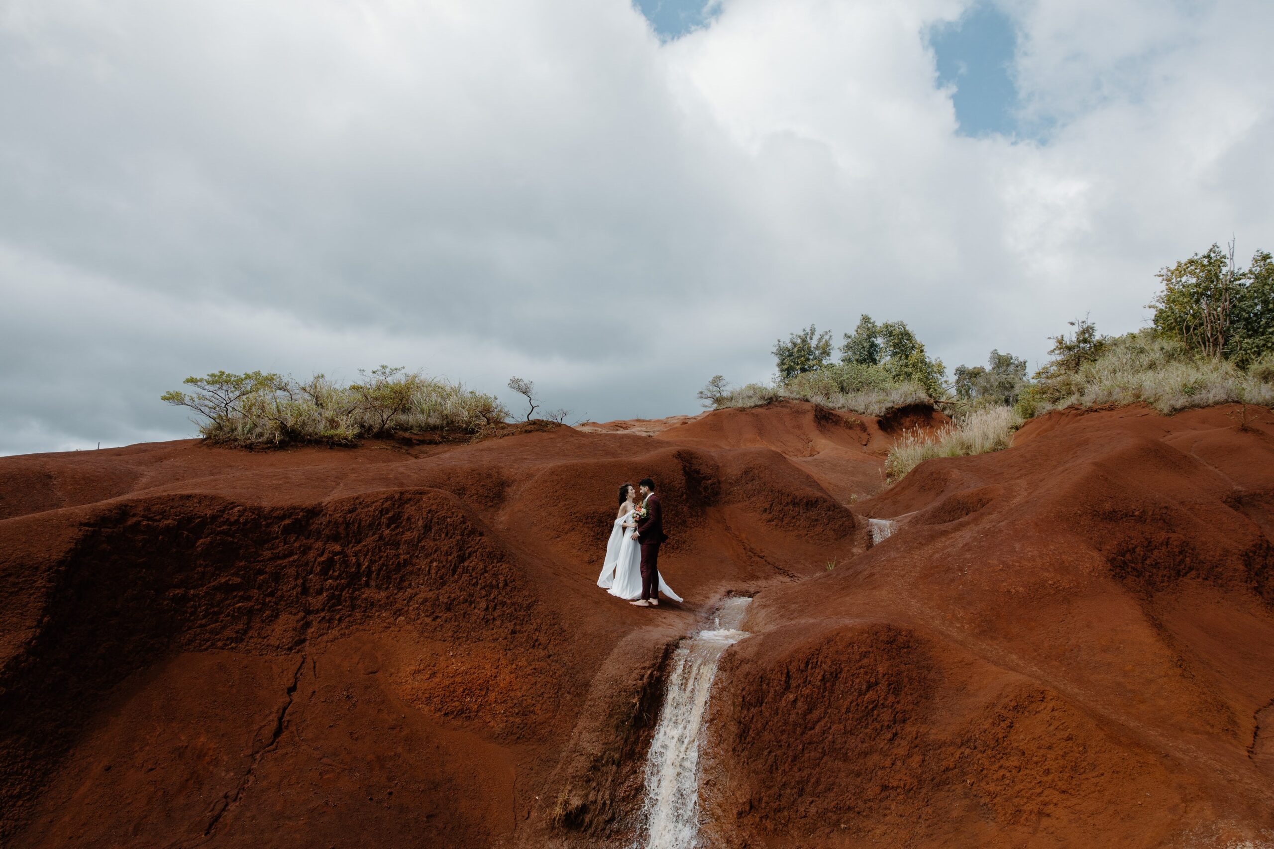 A couple in wedding attire, standing close to one another as they are surrounded by the red Clay of Waimea canyon during their Hawaii elopement